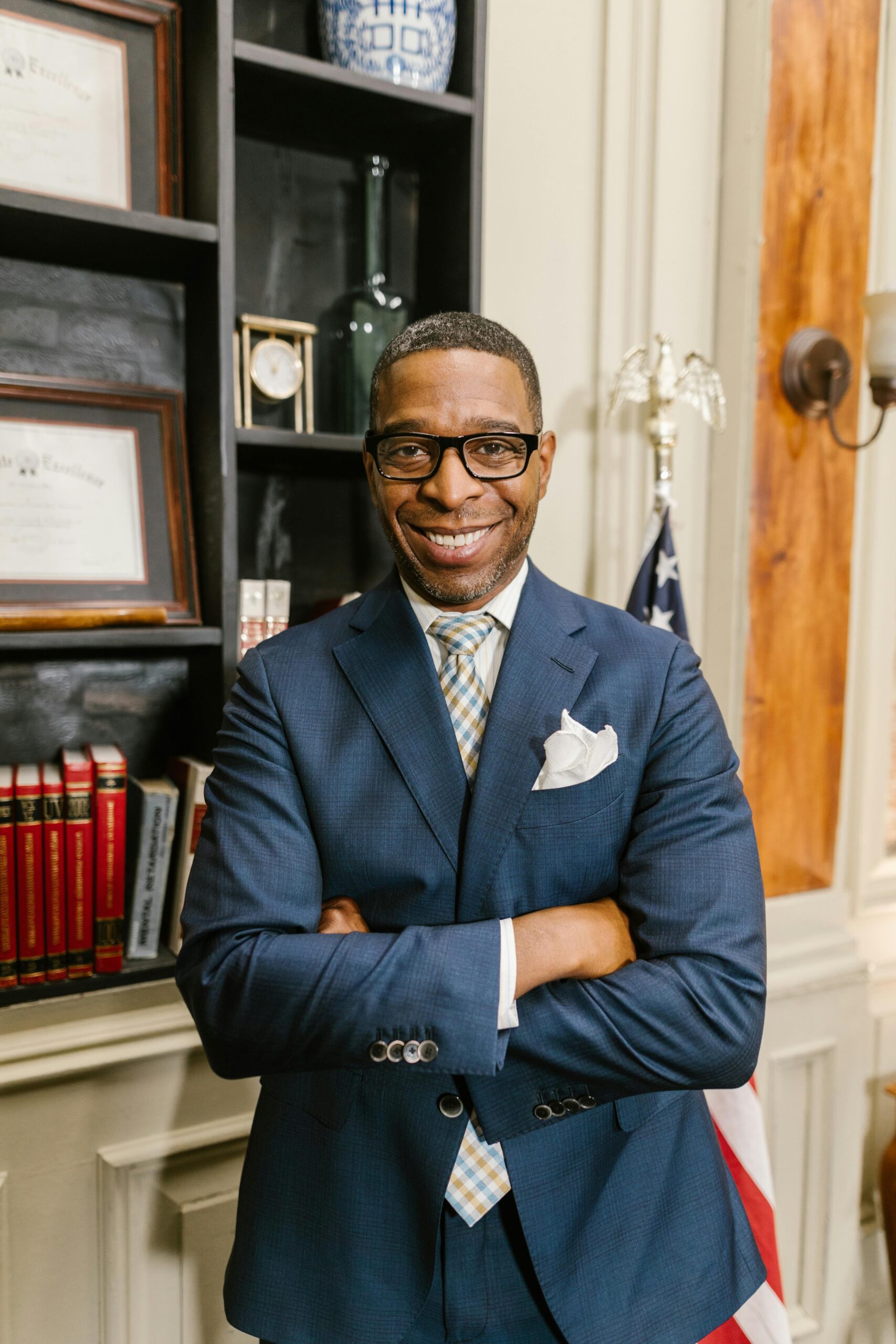 Smiling man with glasses standing confidently in an office with arms crossed.
